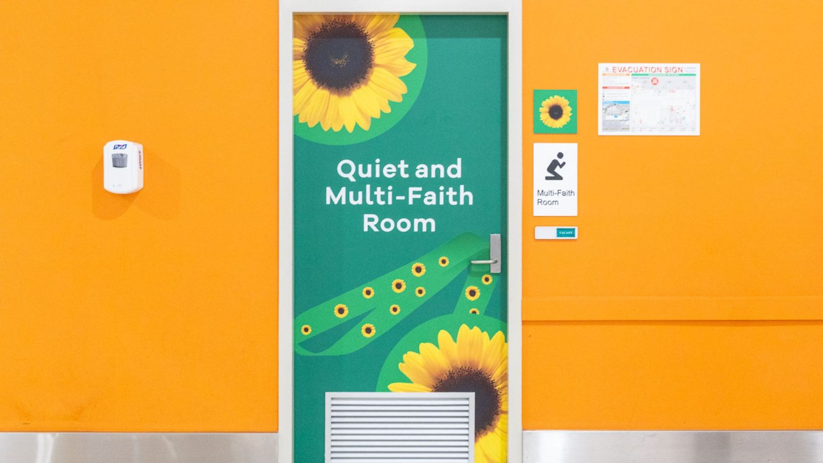 A brightly coloured orange wall featuring the entrance to a Quiet and Multi-Faith Room at Gold Coast Airport. The green door is decorated with sunflowers and a Sunflower Lanyard symbol, with accessibility signage and information nearby.<br/>