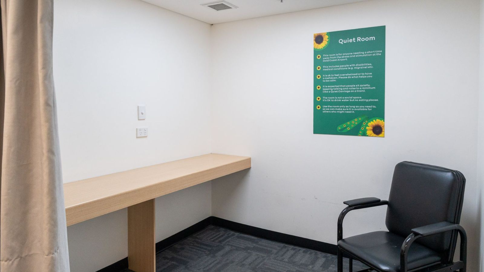 Inside view of a Quiet Room at Gold Coast Airport, featuring a simple wooden bench, a black chair, and a green wall sign with sunflower icons outlining room guidelines. The space is private and softly lit.